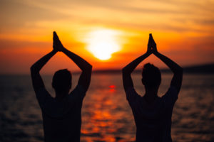 A couple raises their hands in a yoga pose against the light of the setting sun. This could represent the bonds cultivated by working with a Colorado Springs couples therapist. Learn more about online couples counseling in Colorado and other services by searching “couples therapy colorado springs” today. 