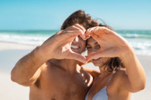 A couple make a heart with their hands while standing on a sunny beach. Learn how a Colorado Springs couples therapist can support your bond with online couples counseling in Colorado today. 